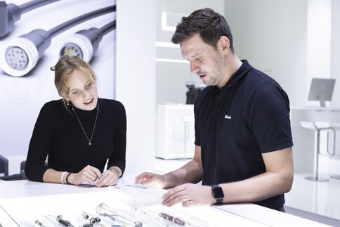 Two people at an exhibition stand bend over a light table on which various technical components are lying.