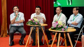 Vier Männer mit Namensschildern diskutieren auf einer Bühne vor rotem Vorhang, Blumen auf Tischen, einer spricht ins Mikrofon, Fokus auf Konferenzatmosphäre.