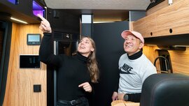 Zwei Personen lächeln in einem modernen Wohnmobil, wobei eine Person auf ein Merkmal an der Decke zeigt.