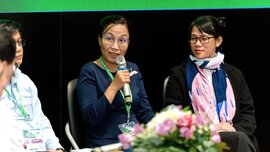 Vier Personen sitzen bei einer Podiumsdiskussion, eine Frau spricht ins Mikrofon. Im Vordergrund steht ein Tisch mit Blumenarrangement.