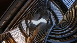 Curved, futuristic building interior with glass and metal, viewed from below towards the bright sky.