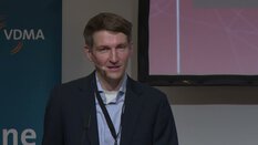 Ein Mann im Anzug spricht mit einem Mikrofon-Headset am Podium. Ein Banner mit 