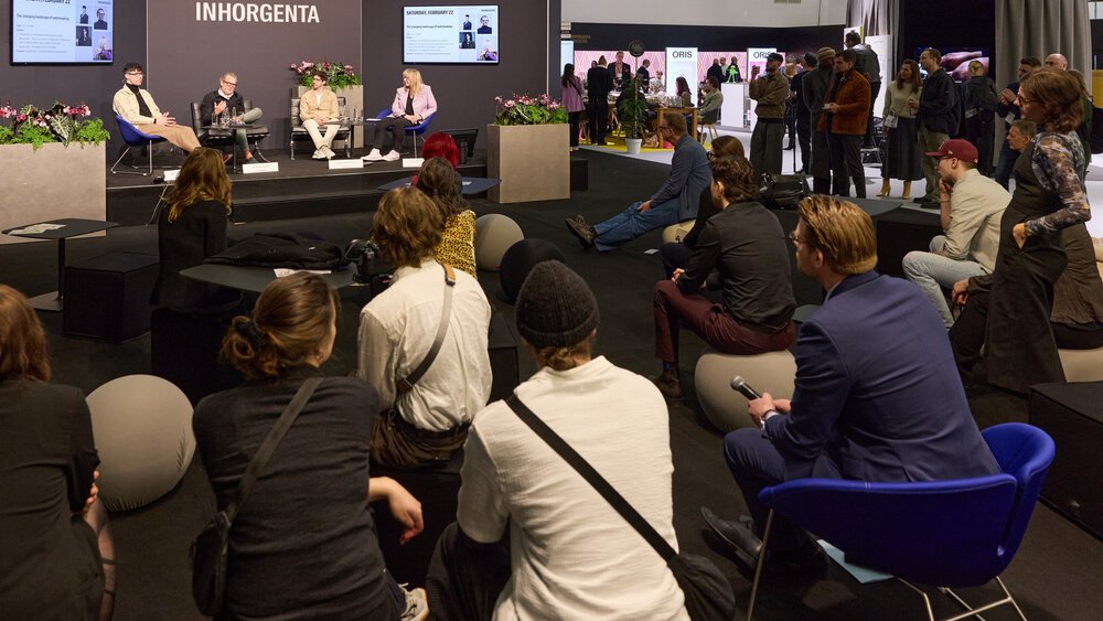 Sitzende und stehende Personen bei einer Podiumsdiskussion auf einer Konferenz mit Präsentationen im Hintergrund.