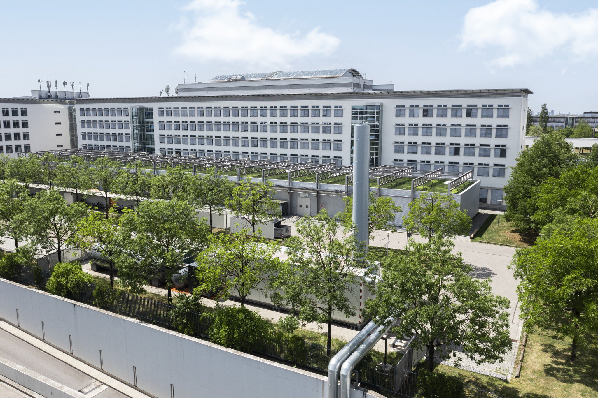 Großes weißes Bürogebäude mit vielen Fenstern, umgeben von Bäumen und Grünflächen unter blauem Himmel.