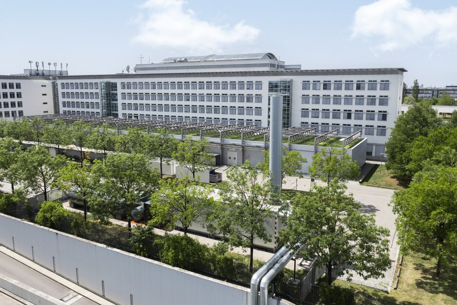 A large white office building with many windows, surrounded by trees and green spaces under a blue sky.