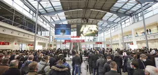 Numerous people stand and wait for admission to productronica. Advertising banners hang from the ceiling.