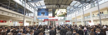 Numerous people stand and wait for admission to productronica. Advertising banners hang from the ceiling.