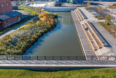 Stadtlandschaft mit rechteckigem Wasserbecken, links dicht bepflanzte Uferzone, rechts Holzstufen und Promenade, umgeben von Gebäuden und Wegen.