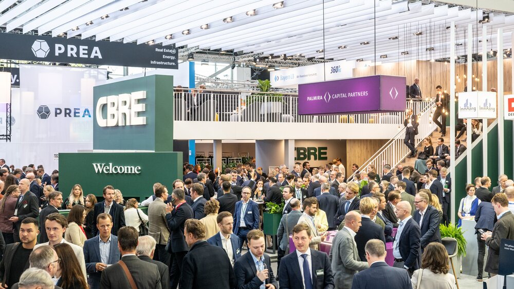 Large crowd making contacts in a brightly lit exhibition hall, with various company stands and signs.