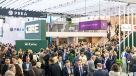 Large crowd making contacts in a brightly lit exhibition hall, with various company stands and signs.