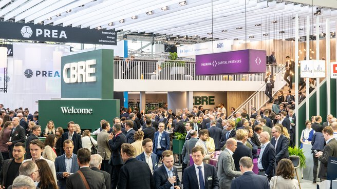 Large crowd making contacts in a brightly lit exhibition hall, with various company stands and signs.