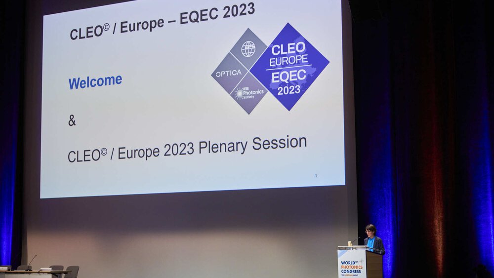 A woman presents on the stage of the World of Photonics Congress next to a large screen on which CLEO Europe logos can be seen.