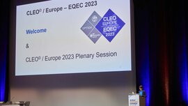 A woman presents on the stage of the World of Photonics Congress next to a large screen on which CLEO Europe logos can be seen.