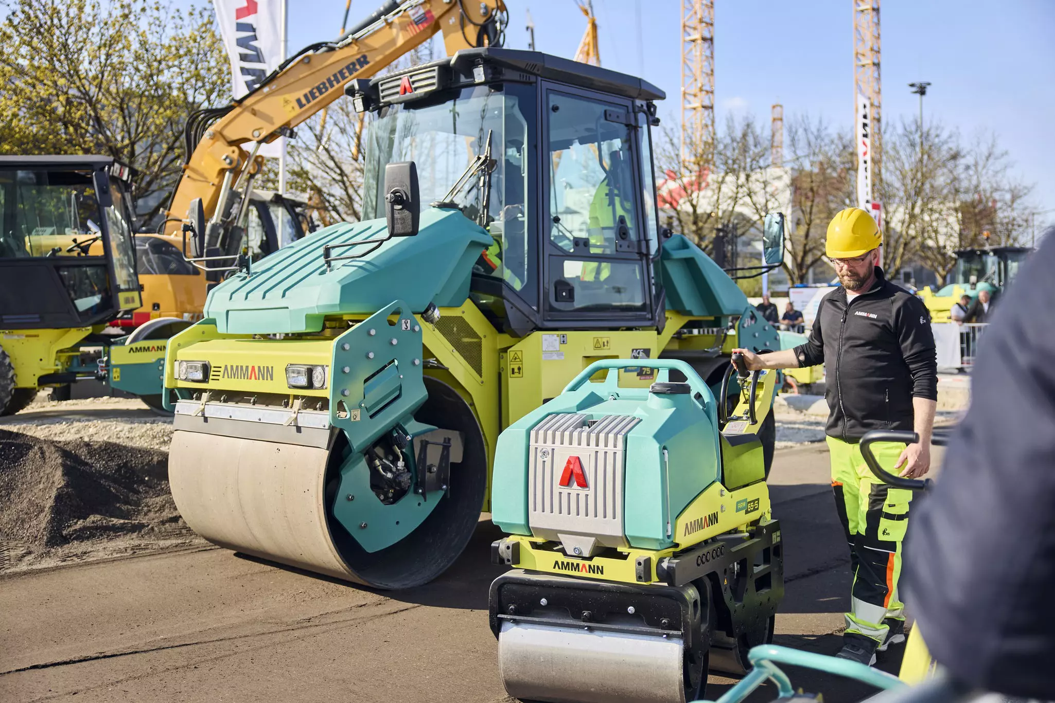 A construction worker operates a small Ammann roller, next to a large road roller at a construction site.
