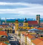 Aerial view of the Munich skyline with historic buildings, under a blue sky with snow-covered mountains in the distance.