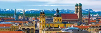 Aerial view of the Munich skyline with historic buildings, under a blue sky with snow-covered mountains in the distance.