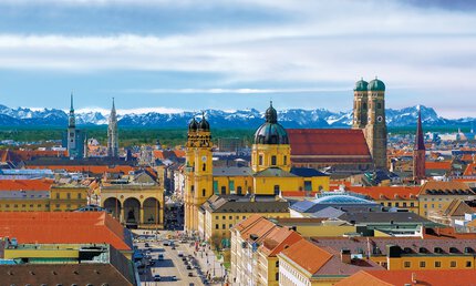 Luftaufnahme der Münchner Skyline mit historischen Gebäuden, unter einem blauen Himmel mit schneebedeckten Bergen in der Ferne.