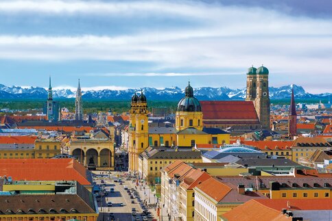 Aerial view of the Munich skyline with historic buildings, under a blue sky with snow-covered mountains in the distance.
