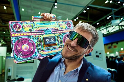 A smiling man with stylish sunglasses is holding a colorful retro-style boombox printed on cardboard on his shoulder.