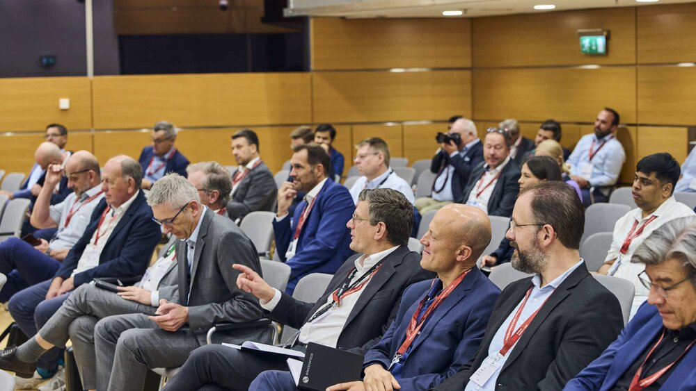 At a conference, people dressed in business attire sit in rows, listen attentively, and take notes.
