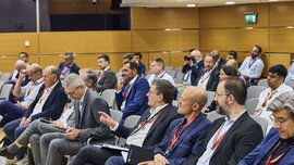 At a conference, people dressed in business attire sit in rows, listen attentively, and take notes.