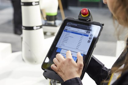 Exhibitor at a trade fair stand holds a tablet for remote control of robotic arms up to the camera.