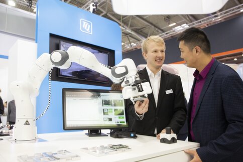 An exhibitor demonstrates a robotic arm with software and remote control to a visitor at his stand. The gripper holds an electronic component.