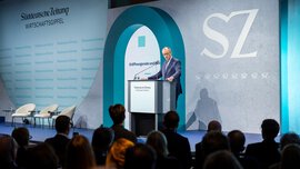 Bundeskanzler Friedrich Merz spricht am Rednerpult auf der Bühne des SZ Wirtschaftsgipfels vor Publikum; im Hintergrund sind das SZ-Logo und die Bühnenkulisse zu sehen. 
