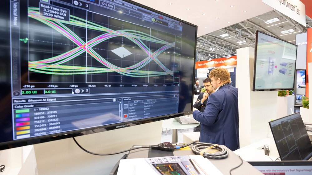 Ein Computermonitor mit bunten Graphen an einem Messestand auf der productronica. Im Hintergrund stehen zwei Männer und betrachten einen Laptopbildschirm.