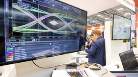 Ein Computermonitor mit bunten Graphen an einem Messestand auf der productronica. Im Hintergrund stehen zwei Männer und betrachten einen Laptopbildschirm.