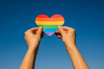 Zwei Hände halten vor einem klaren blauen Himmel ein regenbogengestreiftes Herz und symbolisieren damit den LGBTQ+-Stolz.