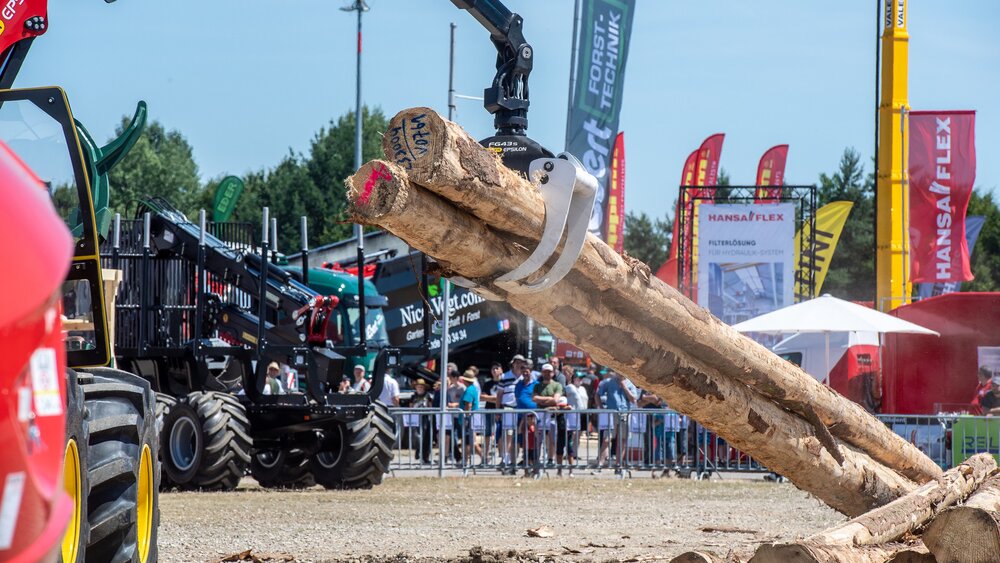 A red construction machine is using a grab arm to lift tree trunks from a pile, whilst other construction machines and trade fair visitors can be seen in the background.