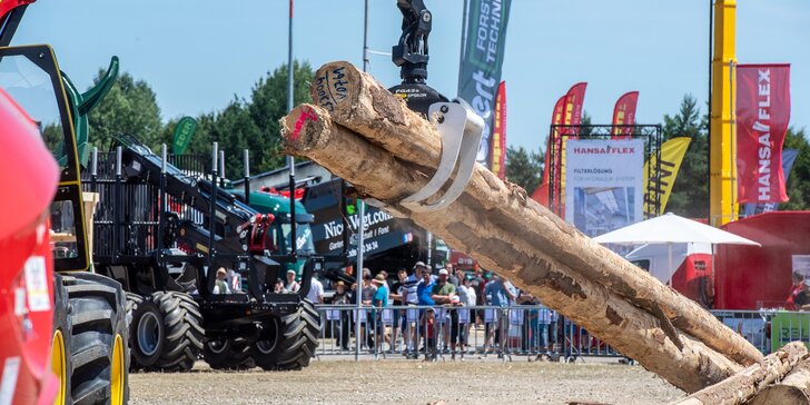 A red construction machine is using a grab arm to lift tree trunks from a pile, whilst other construction machines and trade fair visitors can be seen in the background.