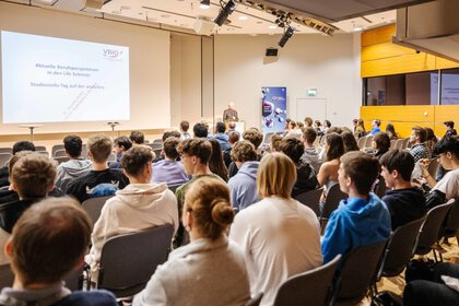  Publikum sieht einem Mann mit Sakko dabei zu, wie er einen Vortrag hält