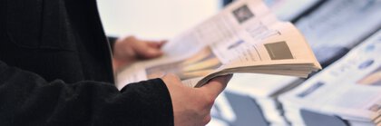 Person in einer schwarzen Jacke hält eine  Zeitung in den Händen und steht vor einer Auslage mit weiteren Zeitungen.