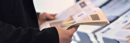 A person in a black jacket holds a newspaper in his hands and stands in front of a shelf with other newspapers.