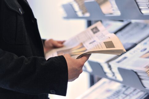 A person in a black jacket holds a newspaper in his hands and stands in front of a shelf with other newspapers.