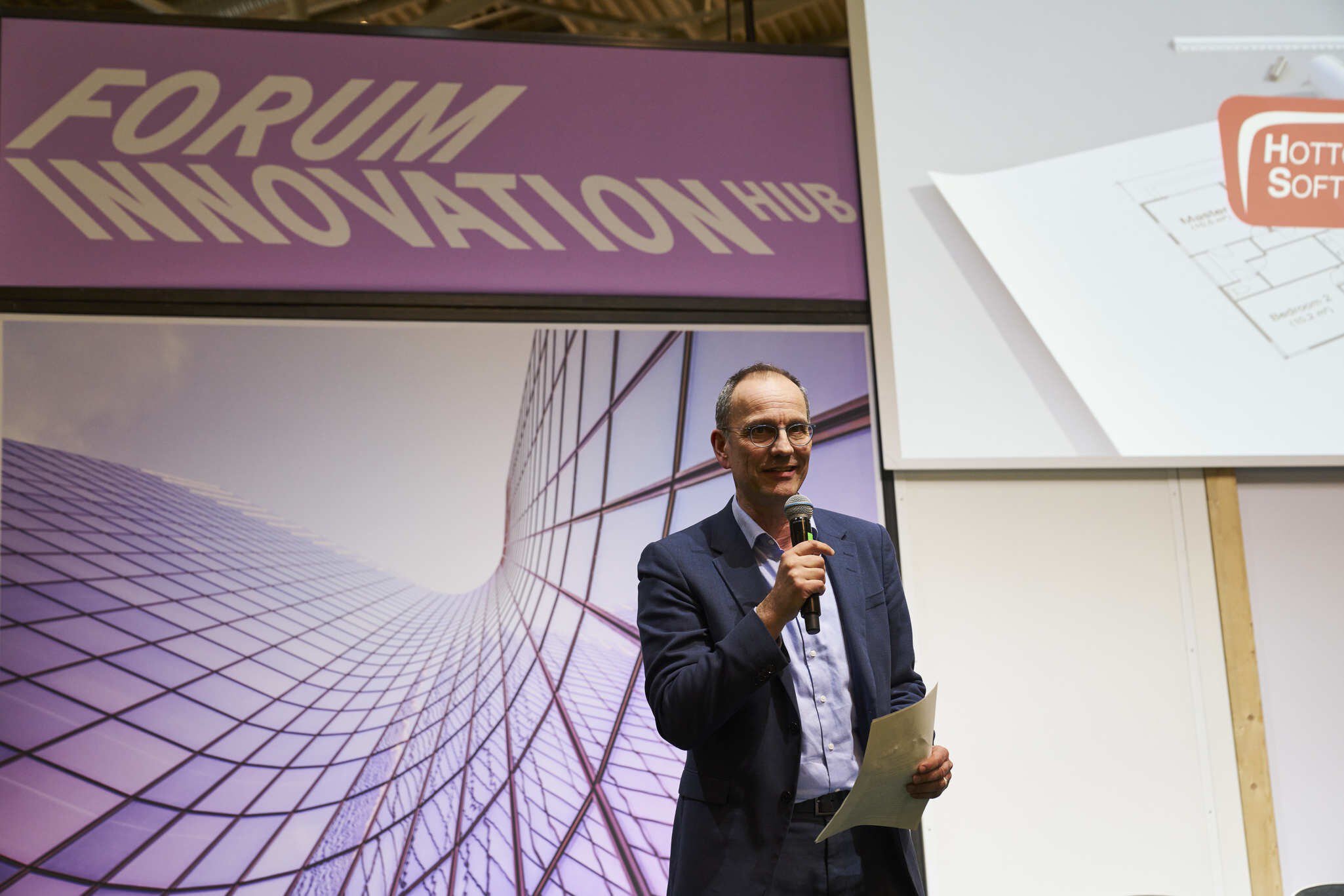 A man wearing glasses and a suit is holding a microphone and speaking into it. Behind him, against a purple background, the words “FORUM INNOVATION HUB” are displayed.
