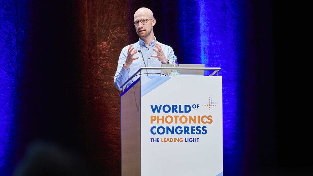 A man in a light blue shirt gestures behind the podium at the World of Photonics Congress.