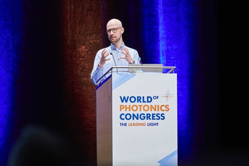 A man in a light blue shirt gestures behind the podium at the World of Photonics Congress.