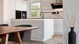 Moderne Küche mit weißen Schränken, Insel, schwarzen Armaturen, Esstisch aus Holz und einer Vase mit Stäbchen.