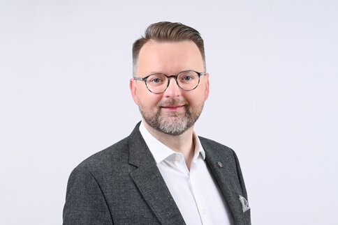 Ein Mann mit Brille und Bart, der eine graue Jacke und ein weißes Hemd trägt, lächelt vor einem schlichten Hintergrund.