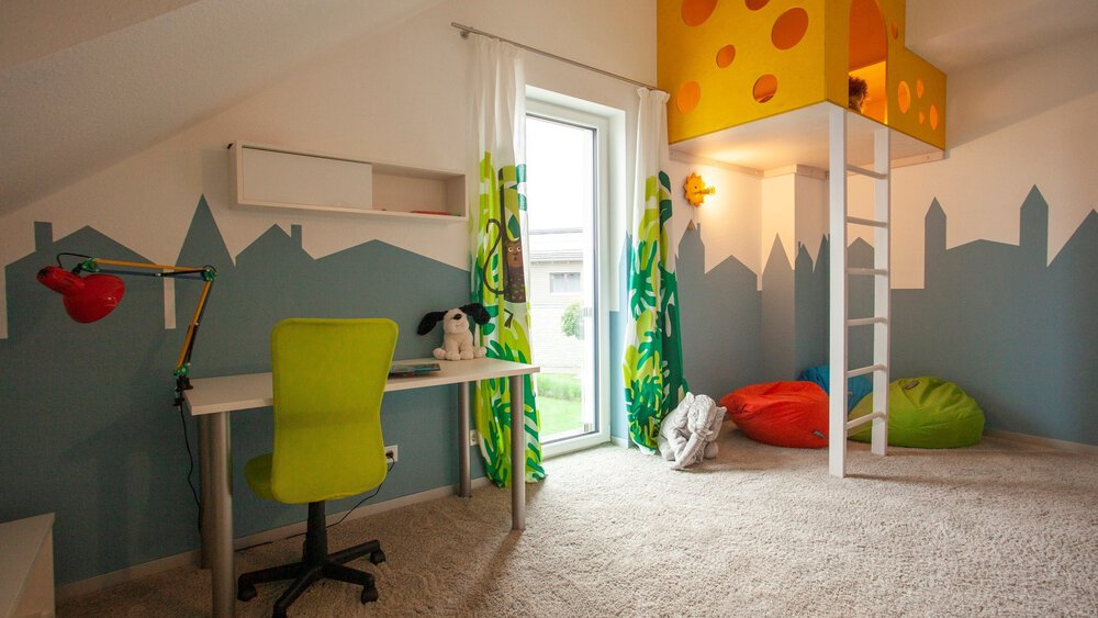 Kinderzimmer mit Teppichboden und Schreibtisch. In der Ecke ist eine Spielhöhle mit Leiter eingebaut ist, darunter liegen Sitzsäcke.