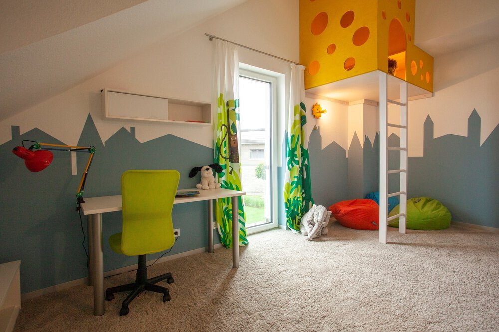Kinderzimmer mit Teppichboden und Schreibtisch. In der Ecke ist eine Spielhöhle mit Leiter eingebaut ist, darunter liegen Sitzsäcke.
