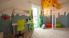 Kinderzimmer mit Teppichboden und Schreibtisch. In der Ecke ist eine Spielhöhle mit Leiter eingebaut ist, darunter liegen Sitzsäcke.