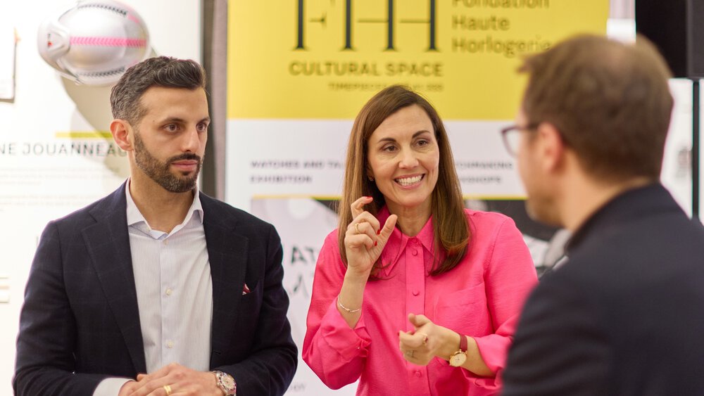 Zwei Personen unterhalten sich mit einem an einem Tisch sitzenden Mann, im Hintergrund ist ein Banner der „Fondation Haute Horlogerie“ zu sehen.
