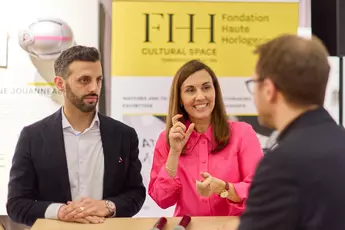 Two people are talking to a man sitting at a table; a banner of the “Fondation Haute Horlogerie” can be seen in the background.