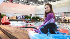Lächelnde Frau im Kajak auf dem Indoor-Paddelbecken der Messe f.re.e, mit Paddel in der Hand und dem Banner „Kanu-Erlebniswelt“ im Hintergrund.