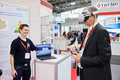 Ein Messebesucher testet eine Virtual-Reality-Anwendung an einem Ausstellerstand, während eine Mitarbeiterin ihn begleitet.