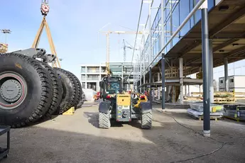 A yellow Liebherr telescopic loader is positioned on a construction site between a modern glass building and large tires being moved by a crane. In the background, construction cranes, scaffolding, and additional machinery are visible.